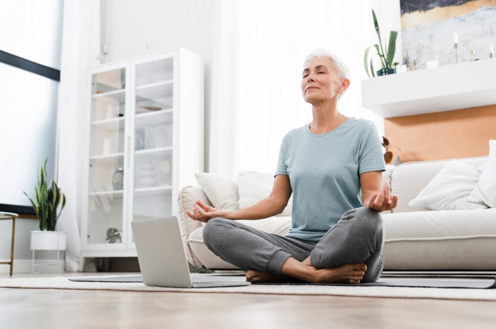 Mature fit athlete meditating in lotus position while watching online training tutorial on laptop.